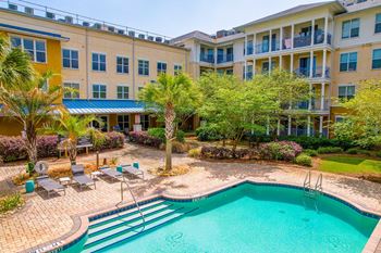 A swimming pool is surrounded by a patio with chairs and a building with balconies.